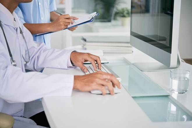 Ein Arzt tippt in einem modernen medizinischen Büro auf einem Computer, während eine Krankenschwester ein Klemmbrett hält.