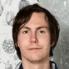 Head and shoulders shot of a young man with dark hair against a patterned wallpaper backdrop.