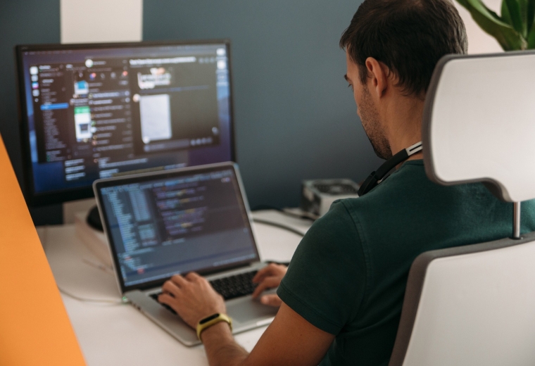 Software developer coding on laptop with dual monitors in modern office workspace.