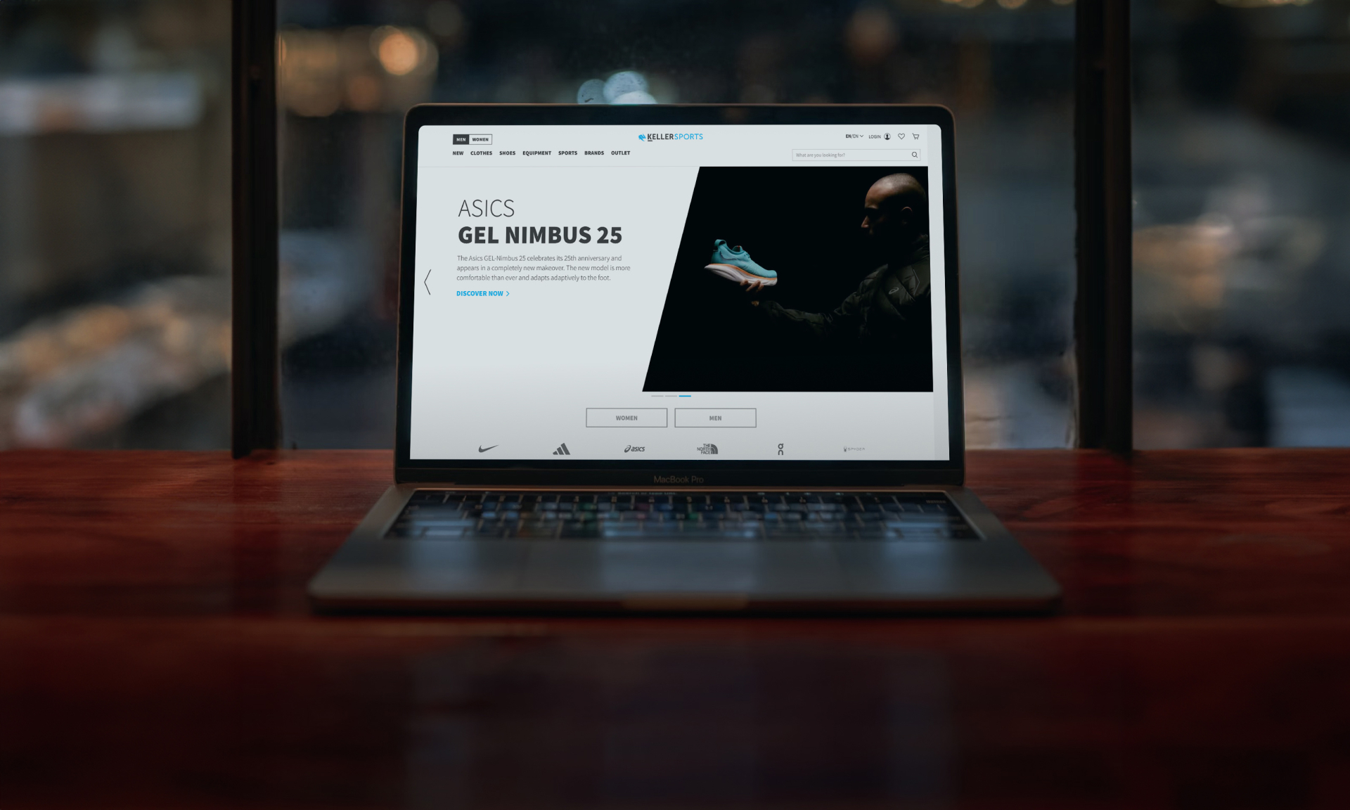 Laptop displaying Asics Gel Nimbus 25 running shoe website on a wooden desk near a window.
