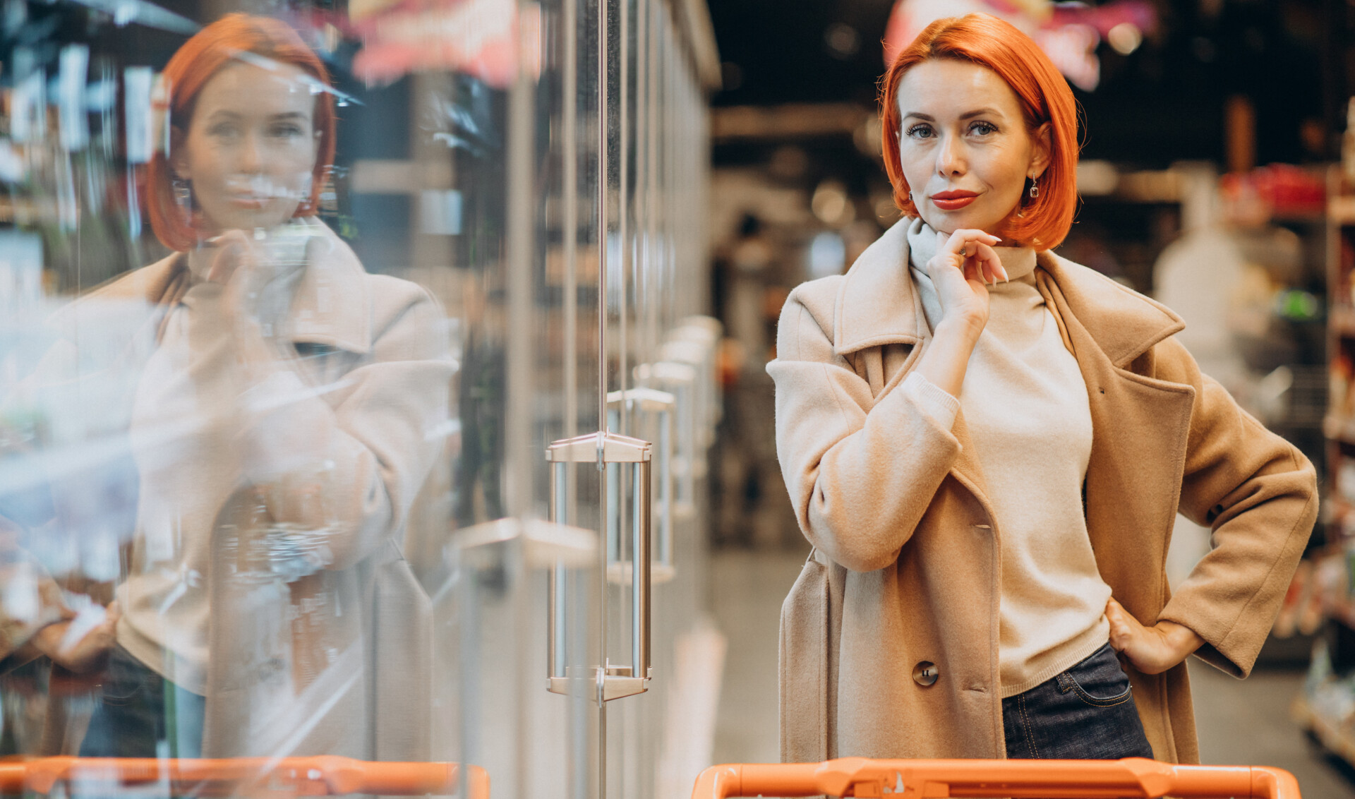 Red-haired woman in a beige coat shopping in a grocery store aisle with a cart, her reflection visible.