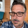 Mann mit Schnurrbart und Brille trägt ein blau kariertes Hemd auf einer Veranda. Porträt.
