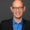 Professionelles Porträt eines lächelnden Mannes mit Brille, blauem Hemd und schwarzem Sakko.