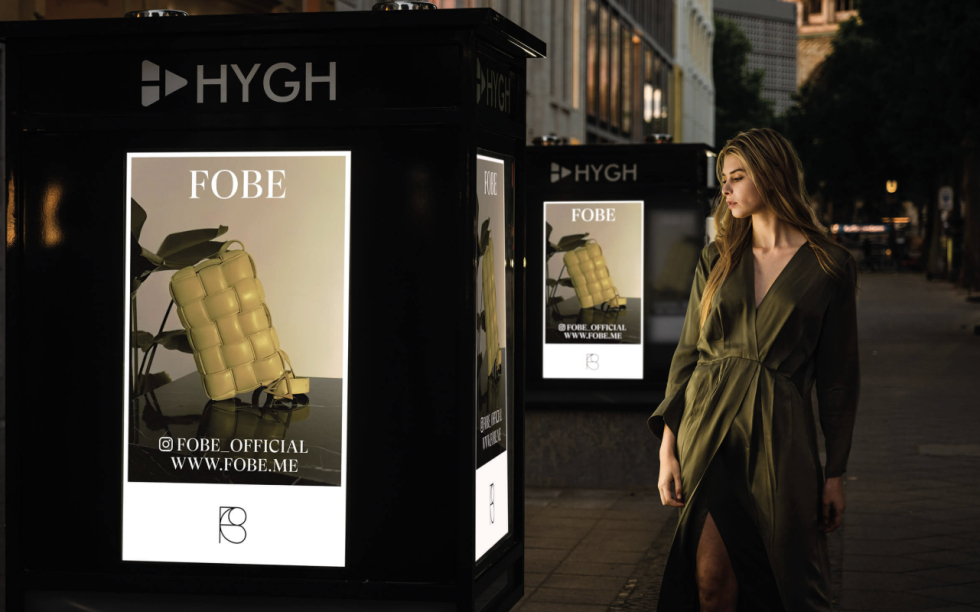 Frau im Kleid steht in der Nähe einer digitalen HYGH-Werbetafel, die für die Handtaschenmarke Fobe wirbt.
