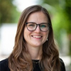Professionelles Porträt einer lächelnden Frau mit Brille und langen braunen Haaren im Freien.