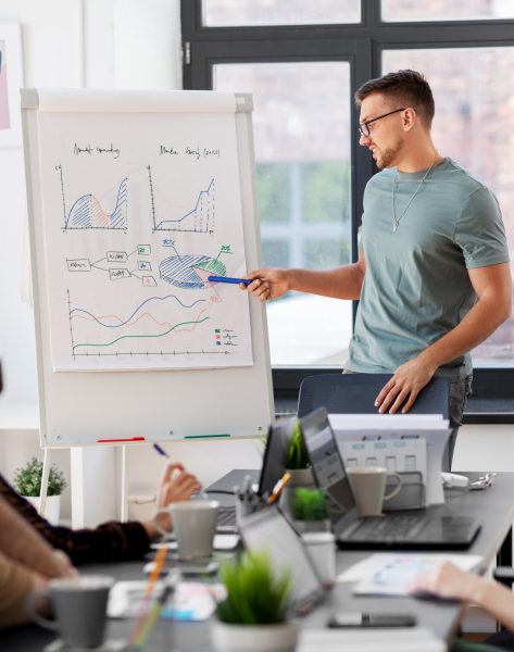 Business meeting: Man presenting data charts on whiteboard to team with laptops.