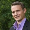 Professional headshot of a smiling man in suit jacket and purple shirt against green foliage. Business portrait.