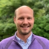 Smiling man portrait; professional head and shoulders shot, wearing a purple jumper, against a backdrop of green foliage.